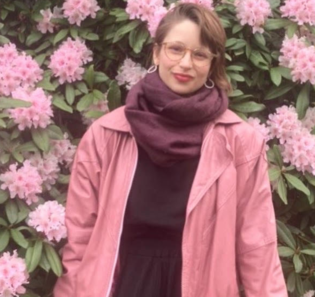 A young woman with brown hair stands smiling in front of a flower bush
